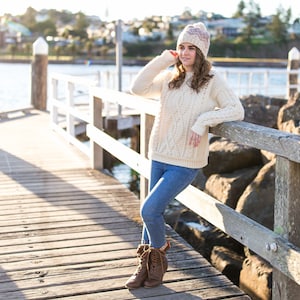 Op de afbeelding: Een vrouw in een witte kabelbreistruien, blauwe jeans en bruine laarzen staat op een houten pier. Ze draagt ​​een roze en witte gebreide beanie en heeft haar hand op haar hoofd. De pier is boven een waterlichaam en er zijn rotsen en bomen op de achtergrond.
