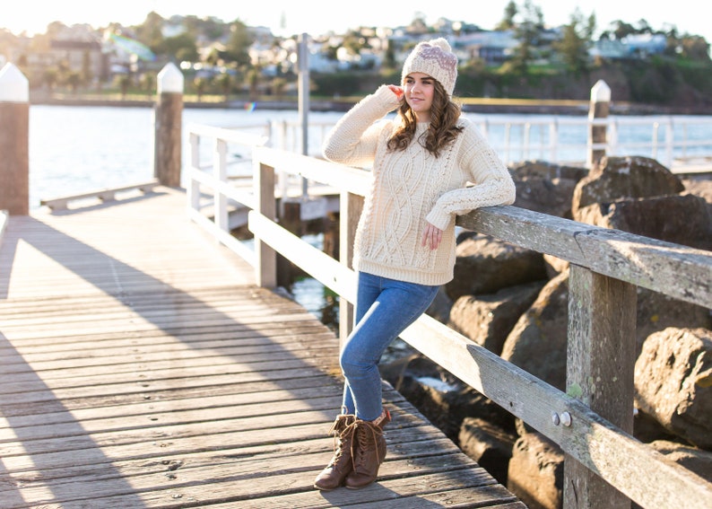 Op de afbeelding: Een vrouw in een witte kabelbreistruien, blauwe jeans en bruine laarzen staat op een houten pier. Ze draagt ​​een roze en witte gebreide beanie en heeft haar hand op haar hoofd. De pier is boven een waterlichaam en er zijn rotsen en bomen op de achtergrond.