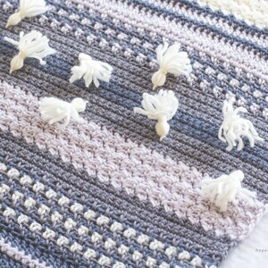 Pode incluir: Uma manta de croch&ecirc; com um padr&atilde;o listrado em tons de cinza, branco e lavanda. A manta &eacute; decorada com pompons brancos.