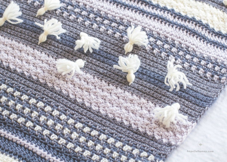 Pode incluir: Uma manta de croch&ecirc; com um padr&atilde;o listrado em tons de cinza, branco e lavanda. A manta &eacute; decorada com pompons brancos.