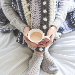 Pode incluir: Uma pessoa vestindo um su&eacute;ter cinza e uma manta de croch&ecirc; listrada, segurando uma caneca branca de chocolate quente. A pessoa est&aacute; sentada em uma cama branca com as pernas cruzadas e usando meias de renda cinza.