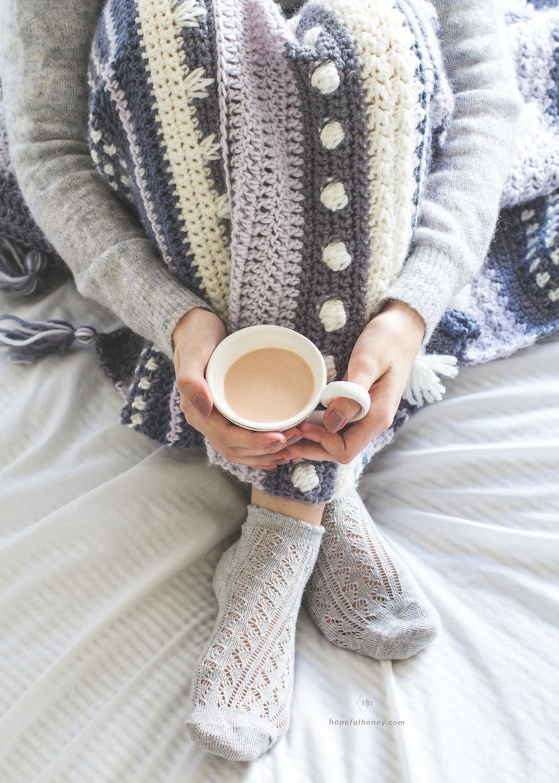Pode incluir: Uma pessoa vestindo um su&eacute;ter cinza e uma manta de croch&ecirc; listrada, segurando uma caneca branca de chocolate quente. A pessoa est&aacute; sentada em uma cama branca com as pernas cruzadas e usando meias de renda cinza.