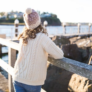 Op de afbeelding: Een vrouw draagt een witte gebreide trui met een kabelbreipatroon en een roze en witte gebreide muts met een witte pompon. Ze staat op een houten pier met uitzicht op het water.