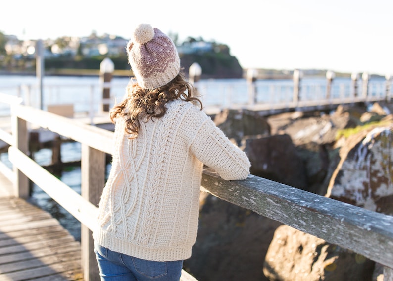 Op de afbeelding: Een vrouw draagt een witte gebreide trui met een kabelbreipatroon en een roze en witte gebreide muts met een witte pompon. Ze staat op een houten pier met uitzicht op het water.