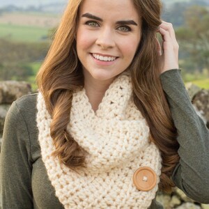 May include: A cream-colored crocheted infinity scarf with two brown wooden buttons. The scarf is worn by a person with long brown hair.