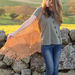 Peut inclure: Une femme portant un gilet en crochet marron avec des franges sur un t-shirt vert et un jean bleu. Le gilet est ouvert &agrave; l'avant et se ferme par des boutons. La femme est debout devant un mur en pierre avec un champ vert et des collines en arri&egrave;re-plan. Le site web "hopefulhoney.com" est visible dans le coin sup&eacute;rieur gauche de l'image.