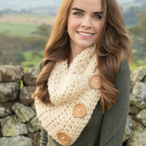 May include: A woman wearing a beige crocheted cowl with wooden buttons. The cowl is worn over a green long-sleeved shirt. The woman is standing in front of a stone wall.