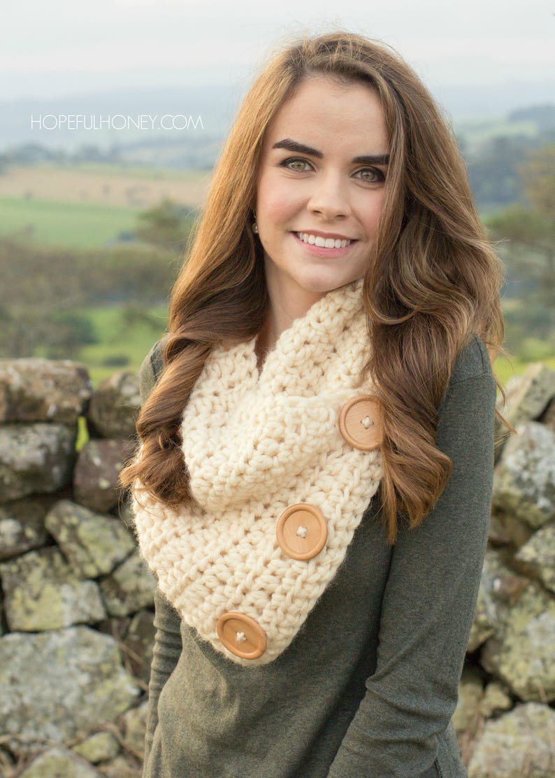 May include: A woman wearing a beige crocheted cowl with wooden buttons. The cowl is worn over a green long-sleeved shirt. The woman is standing in front of a stone wall.