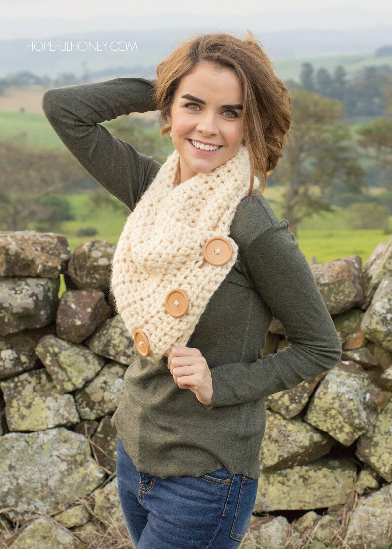 May include: A woman wearing a green long-sleeved shirt and blue jeans is standing in front of a stone wall. She is wearing a crocheted cream-colored infinity scarf with wooden buttons.