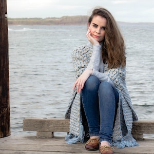 May include: A woman wearing blue jeans and a blue and white crocheted shawl sits on a wooden bench overlooking the ocean.