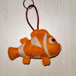May include: Orange and white felt clownfish keychain with a red cord and silver metal ring. The fish has black eyes and white stripes. The keychain is a handmade item.