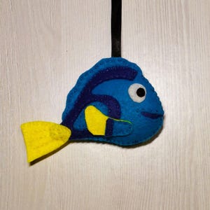 May include: A handmade felt fish keychain in shades of blue and yellow. The fish has a dark blue outline, a yellow tail fin, and a black strap with a silver key ring. The keychain is designed to resemble a cartoon fish.