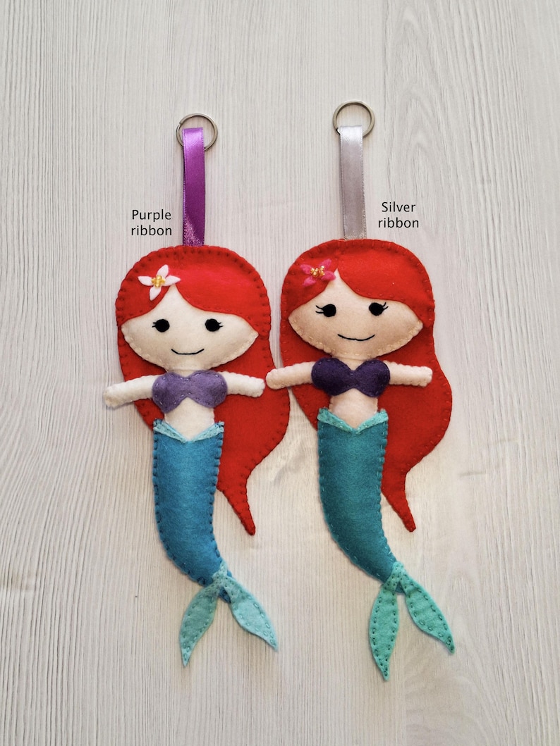 May include: Two felt mermaid keychains with red hair, blue tails, and purple tops. One has a purple ribbon, the other a silver ribbon. Each has a small flower and a metal ring for attaching keys.