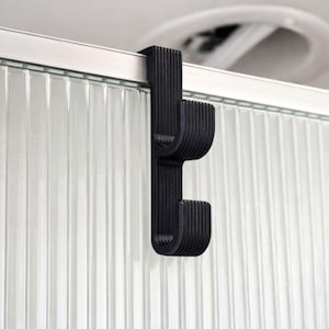 May include: A black over-door hook with a ribbed design. The hook is made of a solid material and is designed to hang over a door or similar structure. The hook is shown against a background of a glass door with vertical lines.
