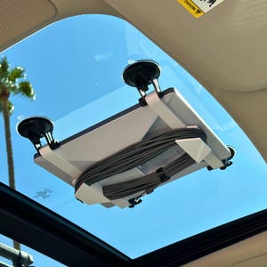 Könnte beinhalten: Ein weißer, aus Kunststoff gefertigter Autohalter mit Saugnäpfen, die an der Innenseite des Schiebedachs eines Autos befestigt sind. Der Halter hält ein Tablet oder ein anderes Gerät. Ein Bündel grauer Kabel ist am Halter befestigt.