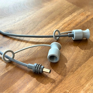 Op de afbeelding: Twee grijze kabeluiteinden met een lus aan elk. De uiteinden zijn gemaakt van flexibel materiaal en hebben een klein gat in het midden. De kabels zijn grijs en hebben een zwarte connector aan het ene uiteinde.