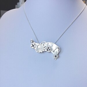 May include: A silver necklace with a pendant featuring a cluster of gears and cogs. The pendant is shaped like a crescent moon and has a dark grey finish.