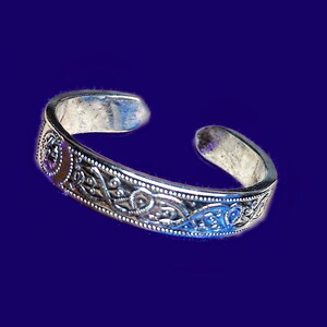 Op de afbeelding: Een zilveren manchetarmband met een ingewikkeld Keltisch knoopontwerp. De armband heeft gedetailleerde patronen en een gepolijste afwerking. Het open ontwerp maakt het gemakkelijk om te dragen. De armband wordt gepresenteerd tegen een donkerblauwe achtergrond.