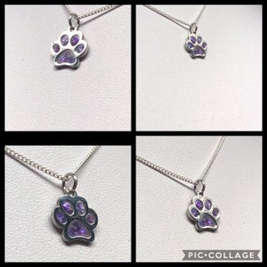 Op de afbeelding: Een zilveren ketting met een hanger in de vorm van een poot afdruk gevuld met paarse glitter. De ketting ligt op een witte achtergrond.