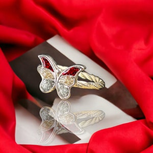 Könnte beinhalten: Ein silberner Schmetterlingsring mit roten und weißen Glitzerakzenten. Der Ring liegt auf einer weißen Oberfläche mit einem roten Satin-Hintergrund.