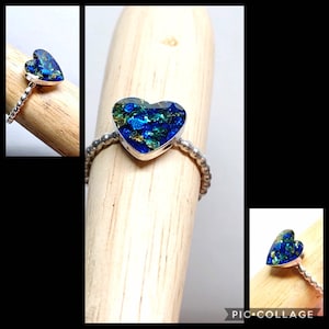 Könnte beinhalten: Ein silberner Ring mit einem herzförmigen, blau-grünen, irisierenden Glasstein. Der Ring liegt auf einer Holzoberfläche.