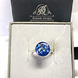 May include: A silver ring with a blue and white resin inlay. The ring is in a white box with a black lid. The lid has the text "Kelandy Designs" in white.