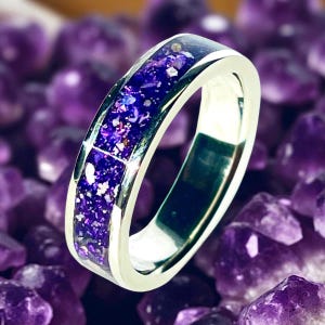 Könnte beinhalten: Ein silberner Ring mit einem Band, das mit einem Mosaik aus tiefvioletten und weißen Fragmenten gefüllt ist. Der Ring ist vor einem Hintergrund aus lila Kristallen platziert, was seine lebendigen Farben und sein einzigartiges Design hervorhebt.