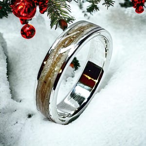 Könnte beinhalten: Ein silberner Ring mit holzähnlichem Inlay und poliertem Finish. Der Einsatz weist hellbraune und cremefarbene Töne auf. Der Ring ist vor einem schneebedeckten Hintergrund mit Weihnachtsschmuck und Grün angeordnet, was auf ein Feiertags-Thema hindeutet.