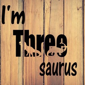 May include: A wooden sign with the text "I'm Three-saurus" in black lettering. The word "Three" has a dinosaur silhouette behind it.