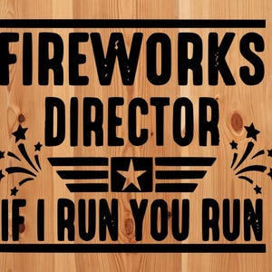 May include: A wooden sign with the text "FIREWORKS DIRECTOR IF I RUN YOU RUN" in black. The design includes a star, a stylized emblem, and starbursts. The background is a light brown wood grain.