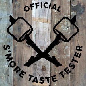 May include: Black graphic with the words "OFFICIAL S'MORE TASTE TESTER" and crossed marshmallow roasting sticks on a rustic wooden background. The design is circular and centered.