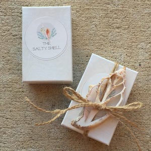 May include: Two white gift boxes with a brown twine bow. The box on the left has a logo that says "The Salty Shell". The box on the right has a seashell tied to the bow.
