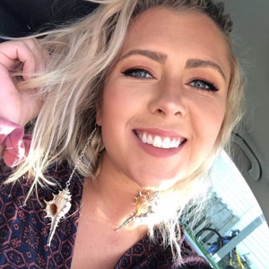 May include: A woman with blonde hair wearing a dark blue and brown patterned shirt with a pair of long shell earrings. The earrings are made of silver chains with a seashell pendant on each side.