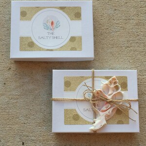 May include: Two white gift boxes with gold polka dot designs. The top box has a logo that says "The Salty Shell" with a watercolor illustration of a seashell. The bottom box is tied with twine and has a seashell on top.