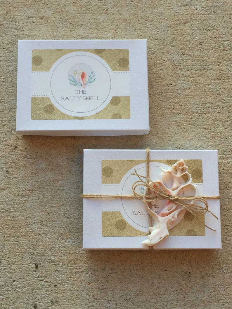 May include: Two white gift boxes with gold polka dot designs. The top box has a logo that says "The Salty Shell" with a watercolor illustration of a seashell. The bottom box is tied with twine and has a seashell on top.
