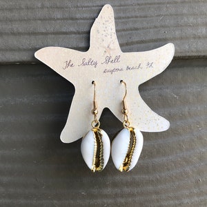 May include: A pair of gold shell earrings with white cowrie shells. The earrings are displayed on a white starfish-shaped card with the text "The Salty Shell Daytona Beach, FL".