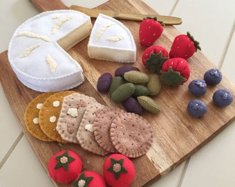 Puede incluir: Una tabla de cortar de madera muestra una rueda de queso de fieltro con una cuña cortada, galletas saladas, aceitunas, fresas, arándanos y tomates. Un pequeño cuchillo de madera descansa en la tabla. Los alimentos de fieltro son de varios colores.