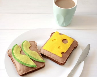 Puede incluir: Dos sándwiches de juguete de fieltro en un plato blanco. Un sándwich está cubierto con una rebanada de queso amarillo de fieltro con agujeros y el otro está cubierto con dos rebanadas de aguacate verde de fieltro. Una taza azul claro con café está en el fondo.