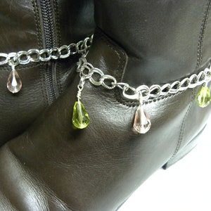 May include: Close-up of brown leather boots adorned with silver-colored chain anklets. Each anklet features two teardrop-shaped beads, one pink and one green. The anklets add a decorative touch to the footwear.