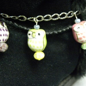 May include: A silver chain anklet featuring three ceramic owl charms. The owls are in purple, green, and pink with white and black details. Each owl has a small bead above and below, with a black background.