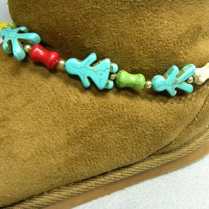 May include: Ankle bracelet with turquoise, red, green, and white beads. The bracelet features small, stylized human figures and other shapes strung together with gold-colored beads. The bracelet is displayed on a brown suede boot.