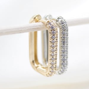 May include: A pair of gold and silver hoop earrings with a rectangular shape and a row of small clear stones around the edge.