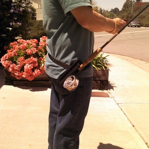 May include: A person wearing a green shirt and blue jeans is holding a fishing rod with a reel attached to their belt. The rod is made of wood and has a black handle.