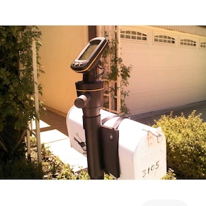 May include: A white mailbox with the number 3105, mounted on a black pole with a GPS device attached. The GPS device has a black screen and yellow accents. The mailbox is in front of a white garage door.