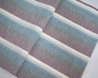 May include: A folded fabric with diagonal stripes in shades of blue, purple, and white. The fabric has a textured appearance, with the stripes running across the material. The overall look is soft and inviting.