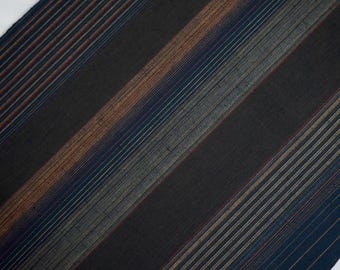 May include: Diagonal view of a textile with a striped pattern. The fabric features a dark background with bands of varying widths and colors, including black, blue, brown, and gray. The stripes run diagonally across the frame.