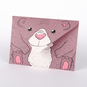 Könnte beinhalten: Ein rosa Umschlag mit einer Cartoon-Illustration eines braunen Bären mit weißem Bauch und rosa Pfoten. Das Gesicht des Bären ist durch den Umschlagklappen sichtbar.