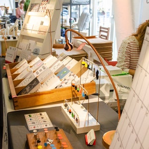 May include: A display of handmade cards, earrings, and other crafts at a market stall. The cards are arranged in wooden display racks. A copper-colored lamp illuminates the items. A calendar is visible in the foreground.