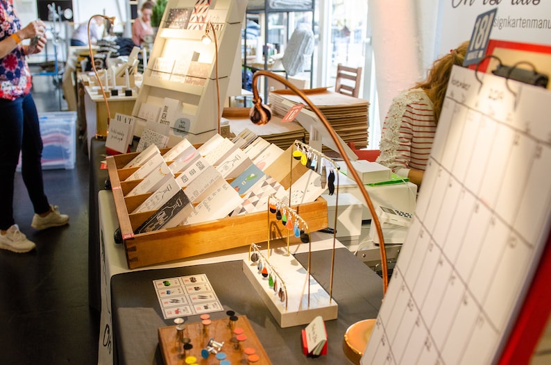 May include: A display of handmade cards, earrings, and other crafts at a market stall. The cards are arranged in wooden display racks. A copper-colored lamp illuminates the items. A calendar is visible in the foreground.