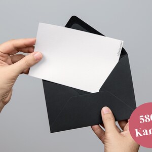 May include: A white card is being removed from a black envelope. The card has text in the upper right corner. A pink circle with the text "580g Karton" is in the lower right corner.